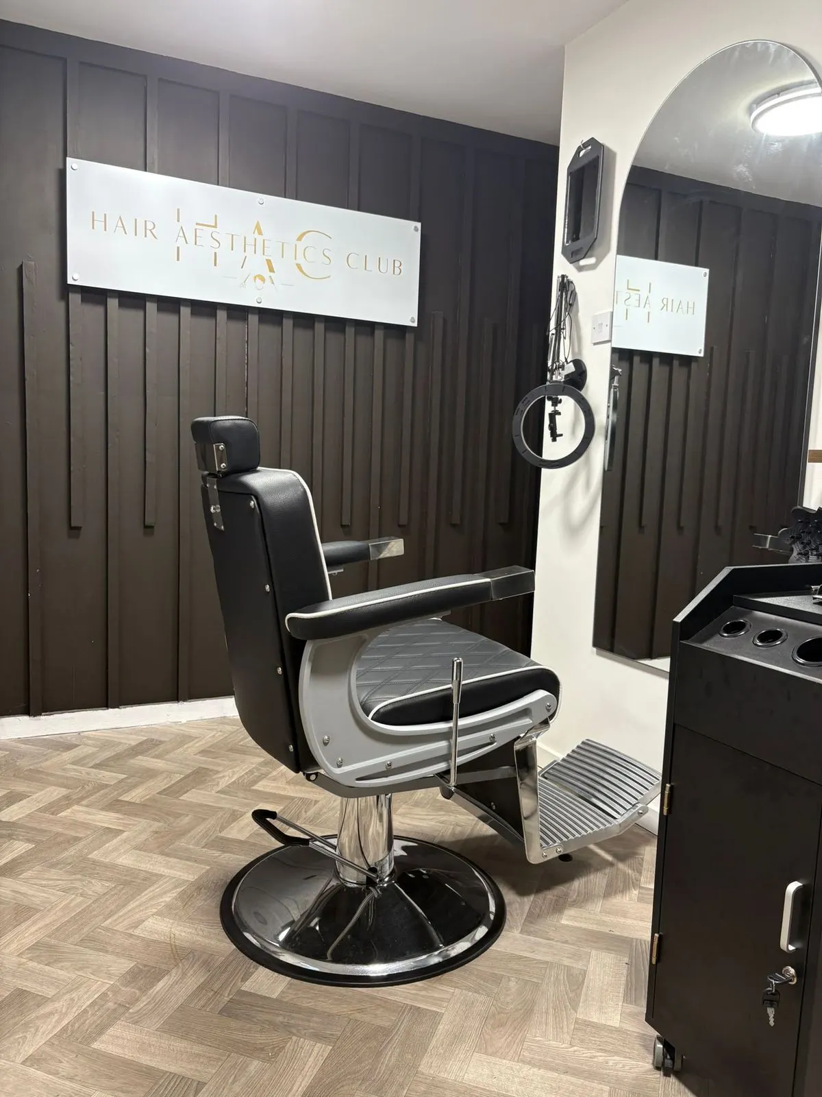 Black-and-chrome barber styling chair beneath the gold Hair Aesthetics Club sign on a dark slatted feature wall, with a herringbone wood floor