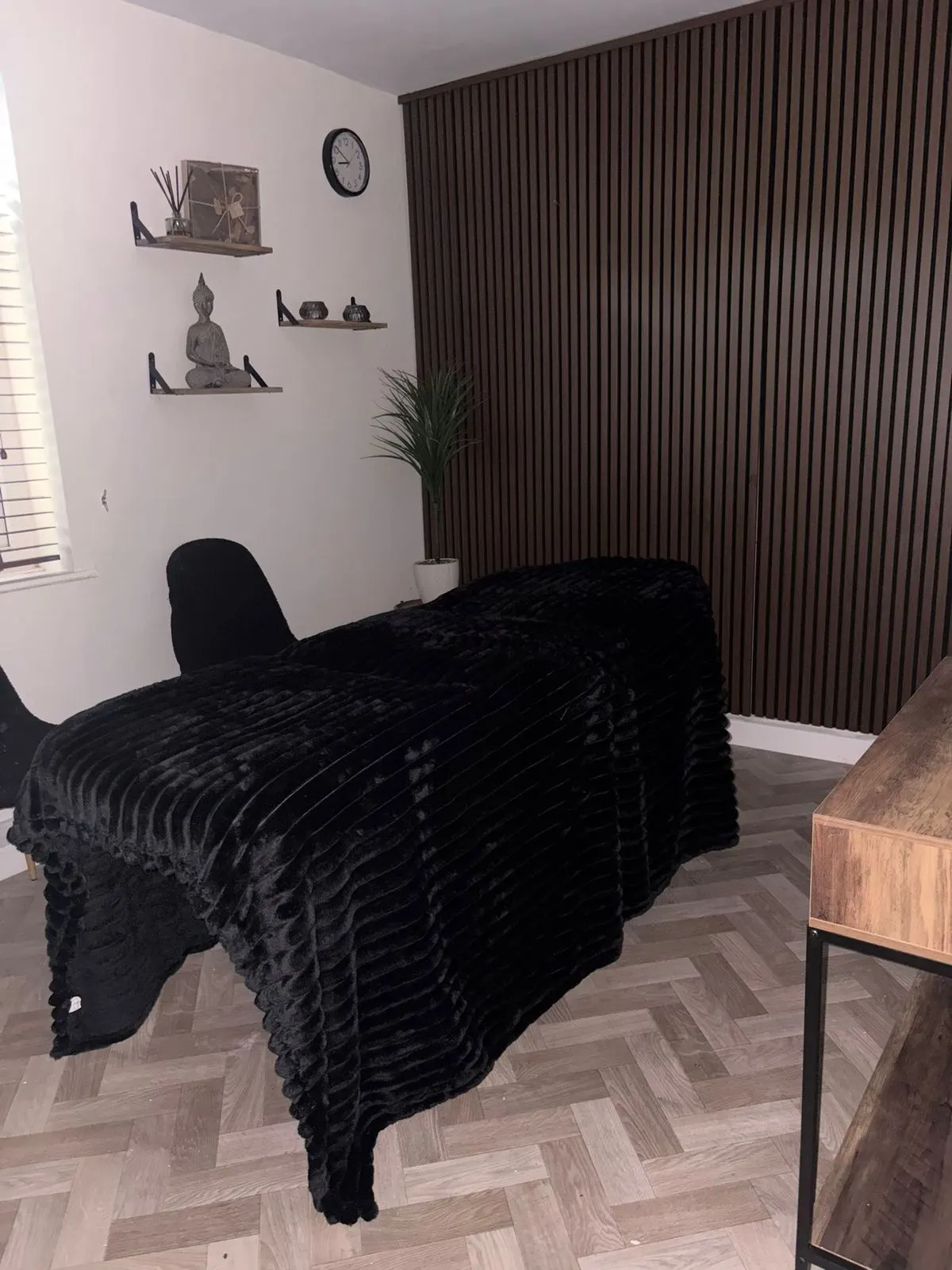 The private treatment room — a therapy couch dressed in black, a walnut slatted wall, shelved decor and calm low lighting